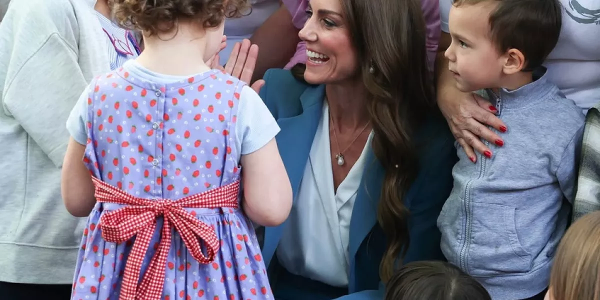 Princess Catherine crouches down to greet young children at an outdoor event, smiling warmly while surrounded by kids and adults.