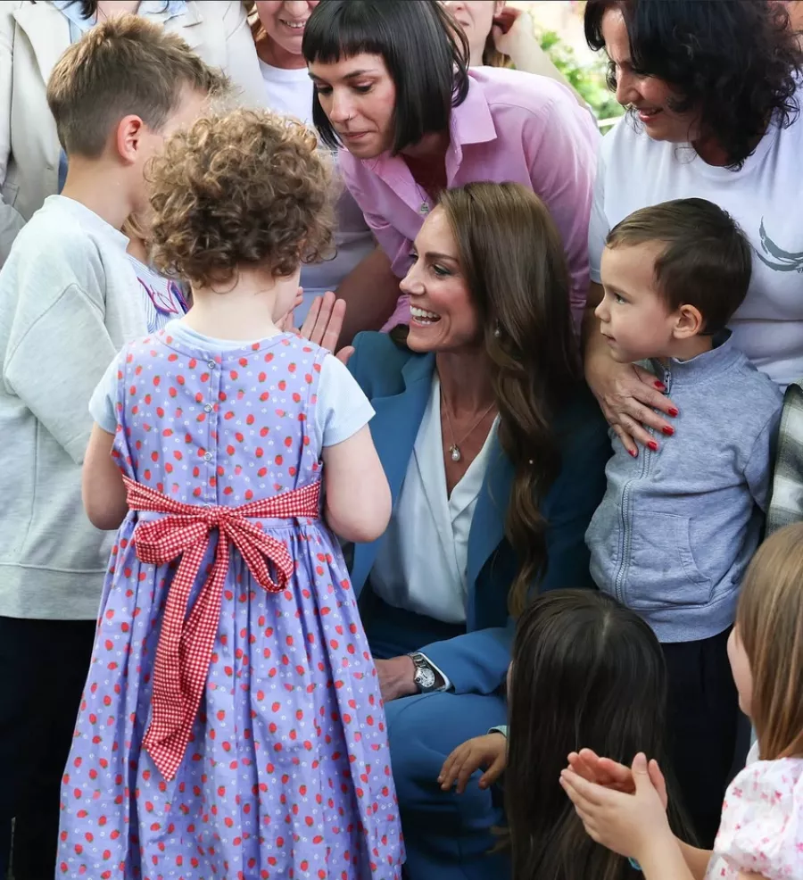 Princess Catherine crouches down to greet young children at an outdoor event, smiling warmly while surrounded by kids and adults.